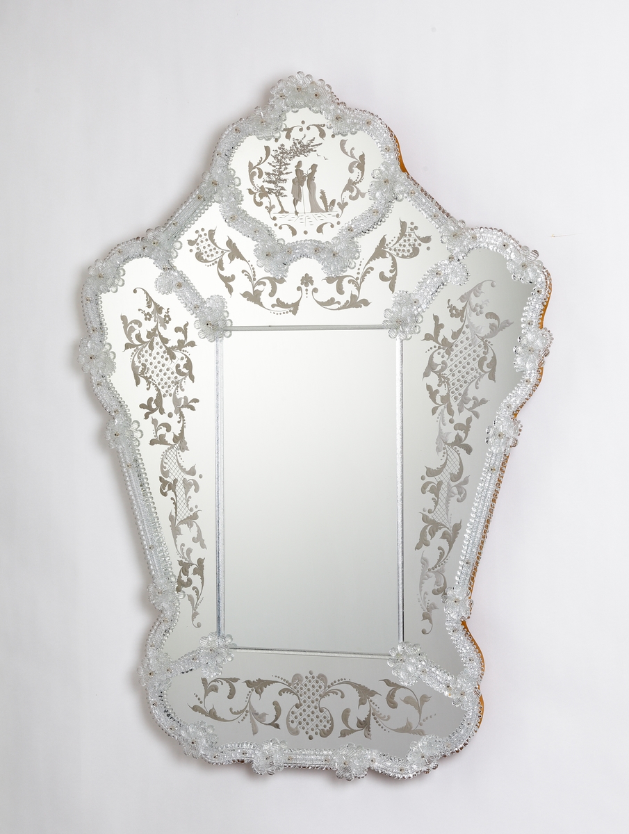 Spegel i venetiansk stil. Ram av skulpterade partier och blommor. Stiliserade graverade växtmotiv runt om, men vid krönet ett kärlekspar i 1700-talsdress.