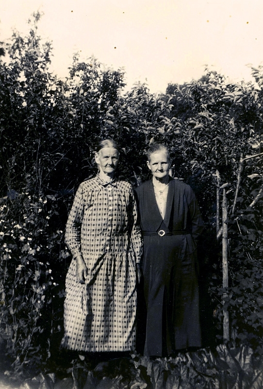 Två systrar står vid en häck, 1920-tal. Från vänster: Maria Henriksson (född Nilsson), Mölnlycke, Råda (1871 - 1936) och Augusta Nilsson gift till Lindbacken, Råda socken (1865 - 1936). Födda i Sporred "Högsåkern". Föräldrar var Johannes Nilsson och Britta Maria Andersdotter. Ytterligare en syster fanns, och det var Sofia "Soffi" Nilsson (1860 - 1945) som var trotjänarinna i Heljered Sörgård "Ordförans".
