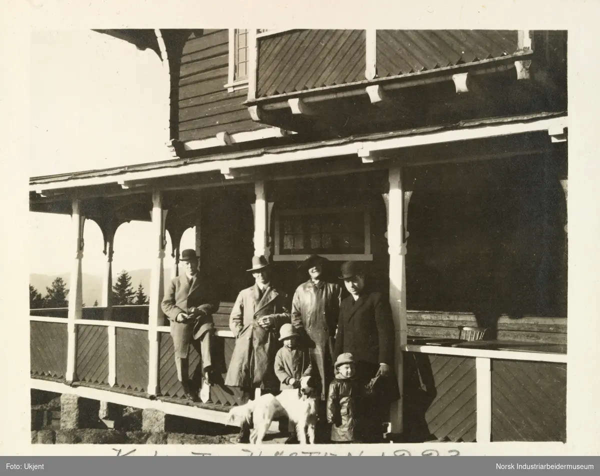 Familie med mann, kvinne, barn og hund foran svalgang på hus. En mann sitter på rekkverk, alle personene har hatt