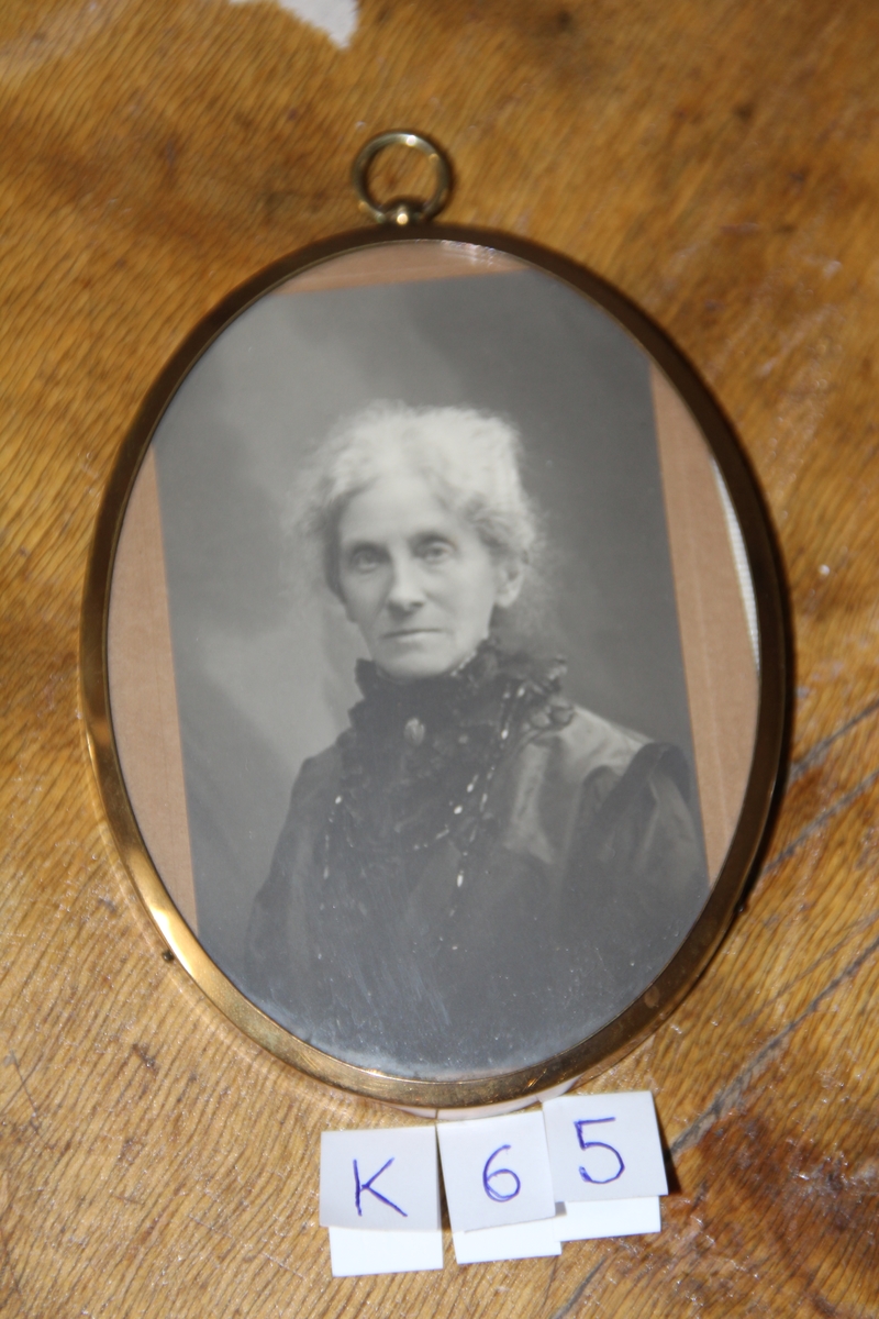 Portrett av eldre kvinne med hvitt hår, venstrevendt, avskåret i livet, Bronsje i halsen og sort perlekjede. Oval smal ramme av metall med blå fløyel på baksiden.
