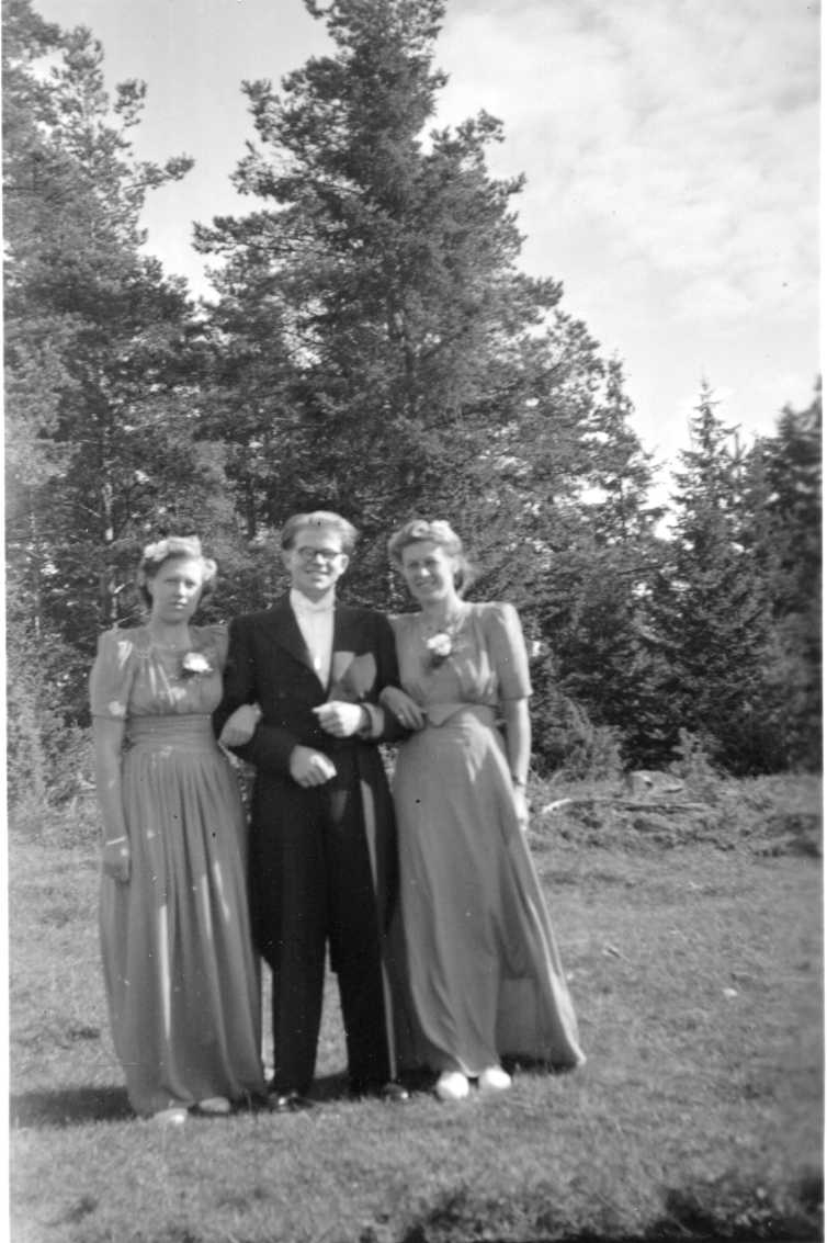 Karl-Erik Johansson i frack stående mellan Ragnhild Wikström till vänster och hennes syster Brita Wikström till höger - Karl-Erik Johanssons hustru. Kvinnorna bär kortärmad långklänning med band i midjan och en ros vid bröstet.