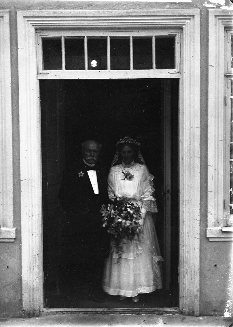 Bröllopsfotografi med Elsa Sofie von Otter och Adam Magnus Emanuel Lagerberg. Hon bär en ljus bröllopsklänning, ett diadem och slöja och håller en blombukett i händerna. Han bär frack. De står sannolikt vid entrén till Ribbagården.