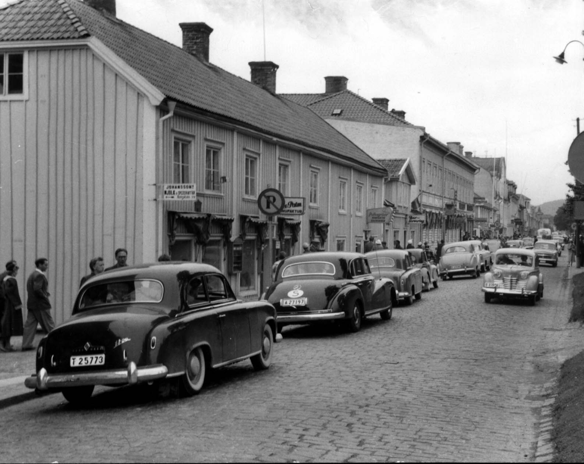 Tät biltrafik på Brahegatan genom Gränna, i raden syns bland annat Mercedes Benz. På husfasaden till vänster skyltas för bland annat Johansson Mjölk & Speceriaffär, Folke Palm Manufaktur och Barservering.