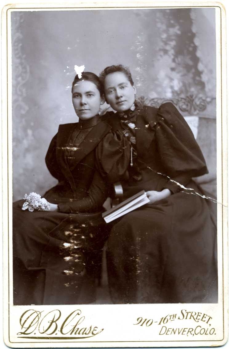 Kabinettsfotografi: gruppbild med två unga kvinnor i mörka klänningar som sitter lutade mot varandra. En kvinna har en blomma i knät och den andra kvinnan håller i en bok.