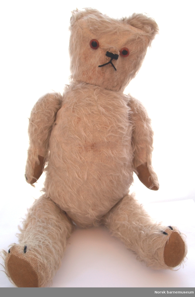 Lys teddybjørn med påsydd brunt stoff. Nese og munn markert med svart tråd. Slitt. knapper som øyne. Kan være nyere enn teddybjørnen.  