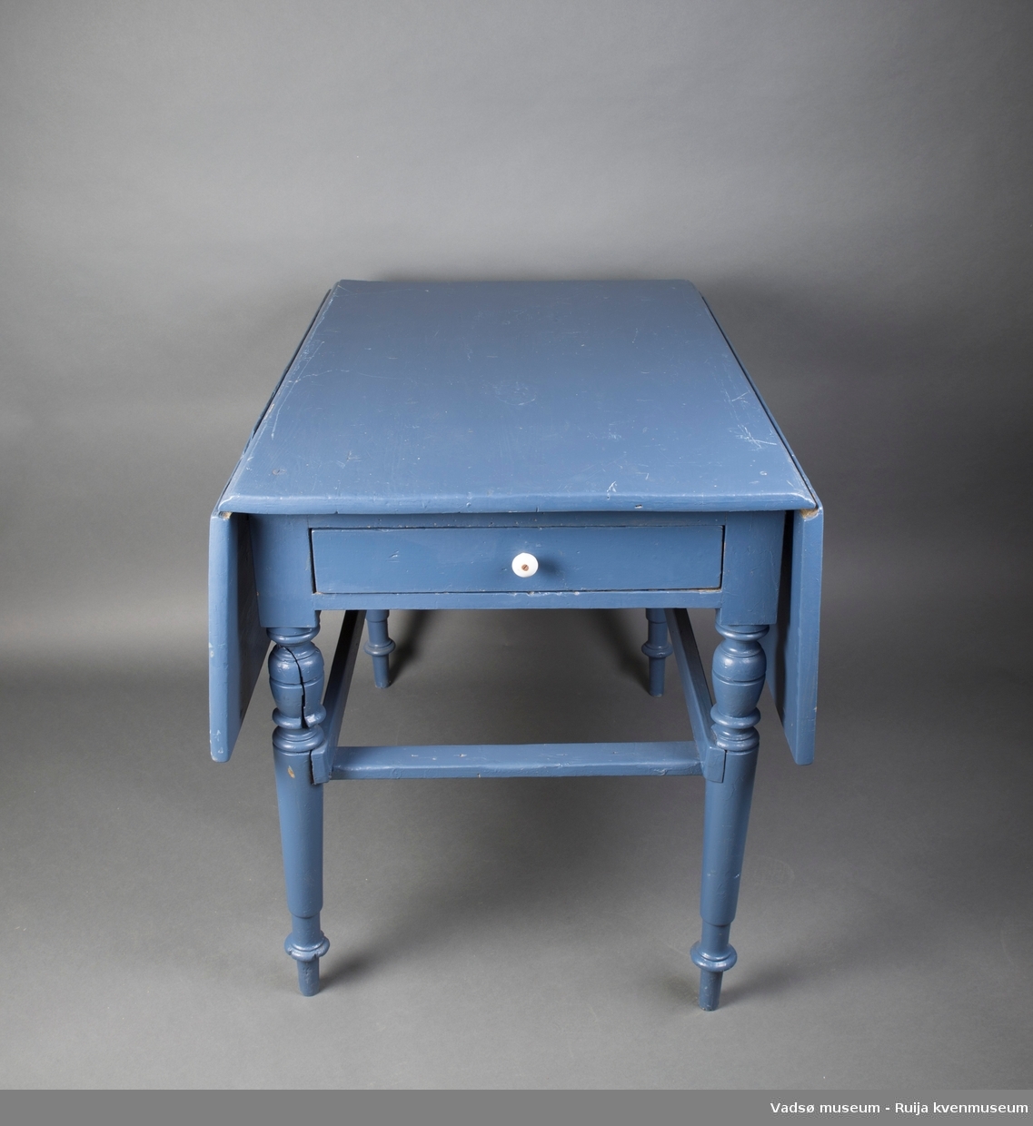Rektangulært trebord med dreide bein. Bordet har hengslede sideklaffer. I ene kortenden er det en skuff med emaljert håndtaksknott. Bordet er malt i blått.