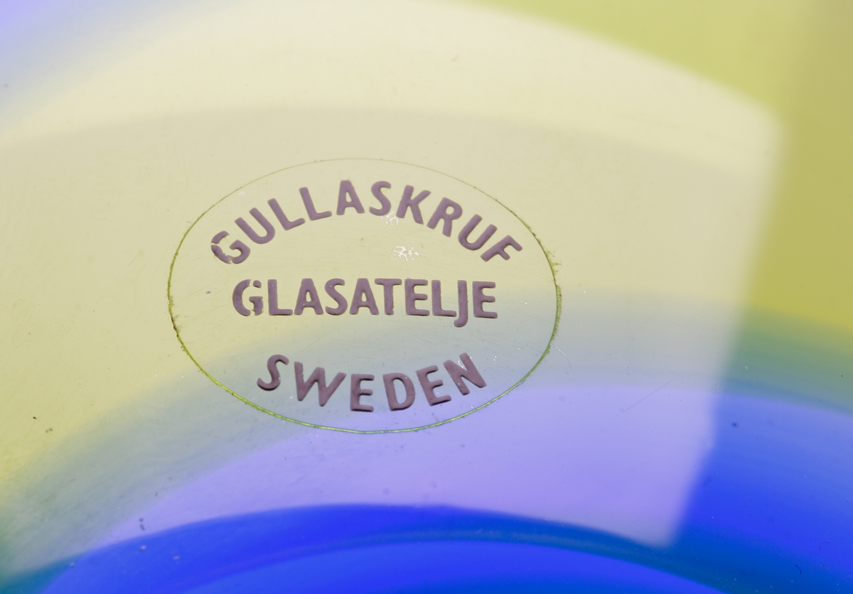 Rund skål färgad med vridna gul-blå linjer.
Etikett. Ofärgad med svart text "Gullaskruf // glasateljé // Sweden"