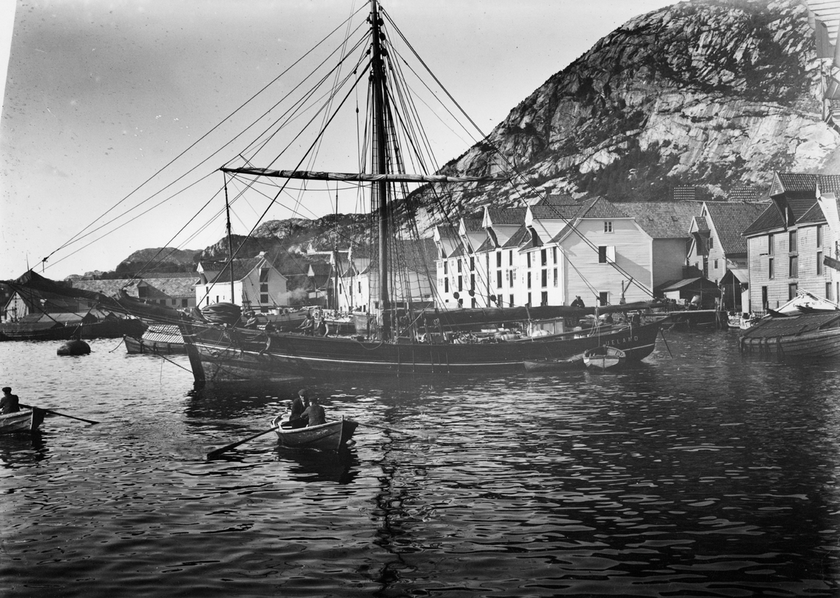 Motivet viser en nordlandsjakten UELAND (bygget 1884) ankret opp utenfor hvite sjøhus i ukjent kystlandskap. Flere menn i arbeid ombord på farkosten. Robåt med to menn fremst i motivet.