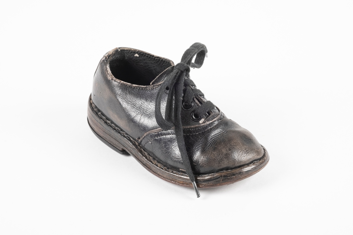 Et randsydd barnesko (høyre sko) av lær. Skoen har snøring med flate lisser. Maljene er av metall. Sålen er av lær og er forsterket med spiker på hælen og ved tuppen på undersiden.