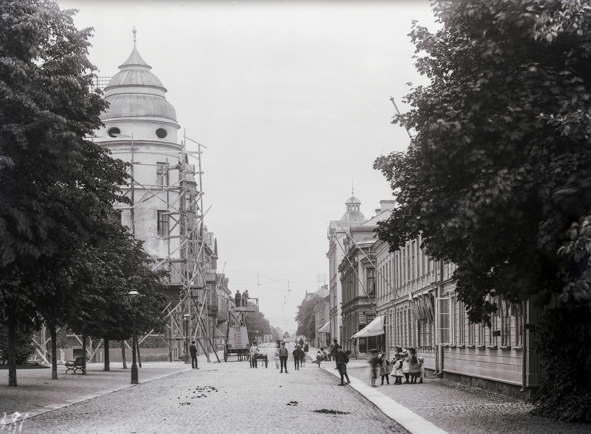 Klostergatan mot söder från Nygatan i Jönköping. Två män står i en hästdragen arbetsvagn och långt bort skymtar en spårvagn. Till vänster ligger sofiakyrkans park. Arkitekt till fastigheten, som omges av byggnadsställning, var August Atterström.