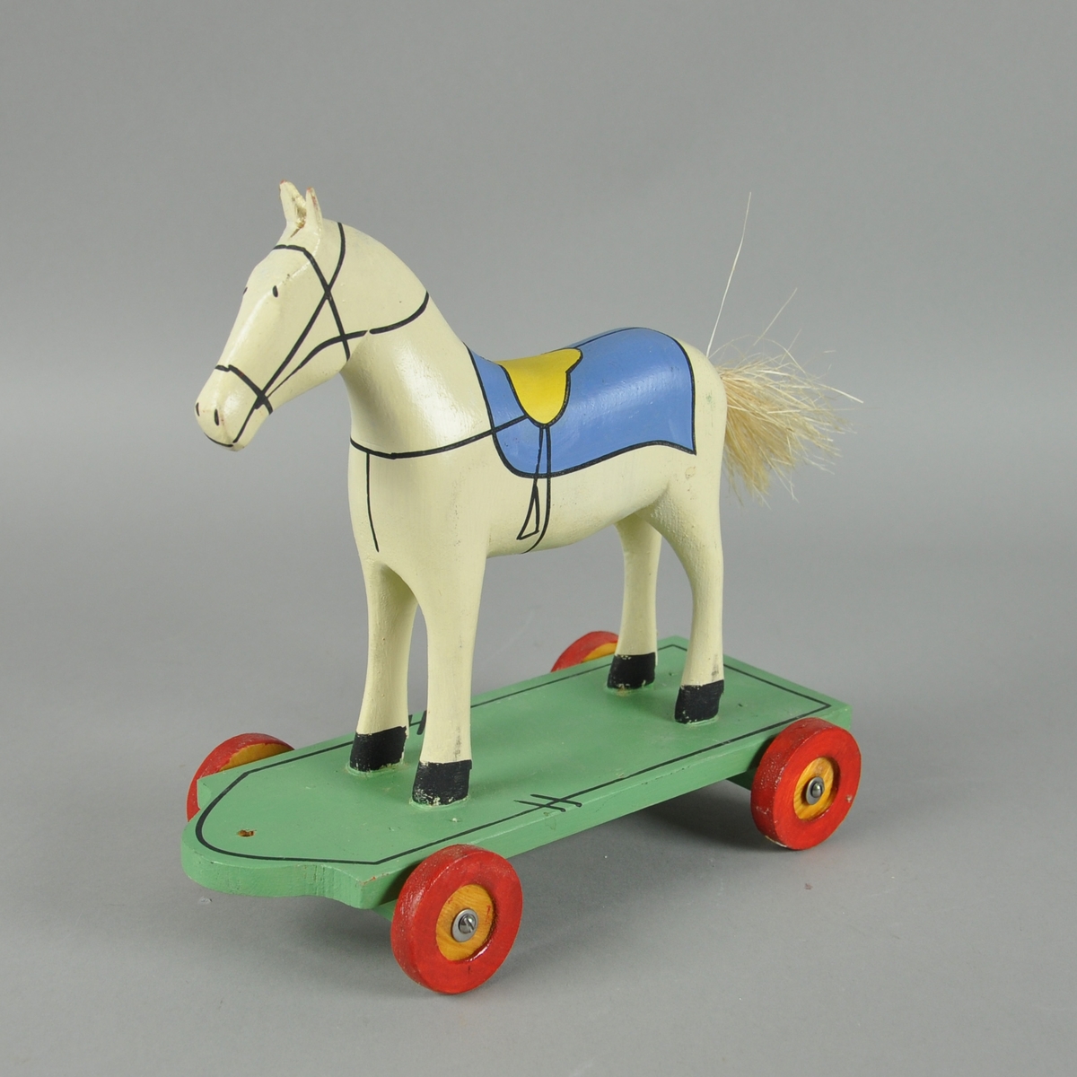 Hvit trehest med påmalt sadel, montert på grønt trebrett med fire hjul. Hesten har hale av bast.