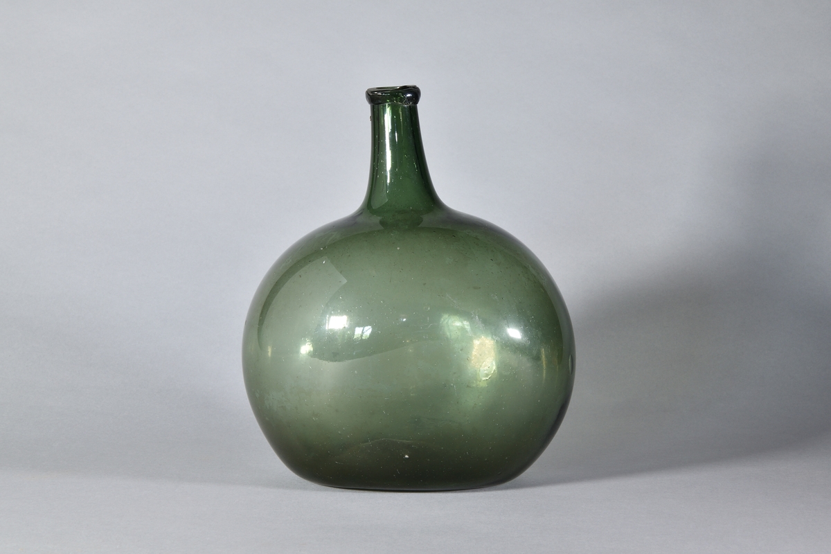 Flaska av handblåst grönt glas, bukig med tillplattade sidor. Hals med pålagd ring kring mynningen, svagt inbuktad i botten.