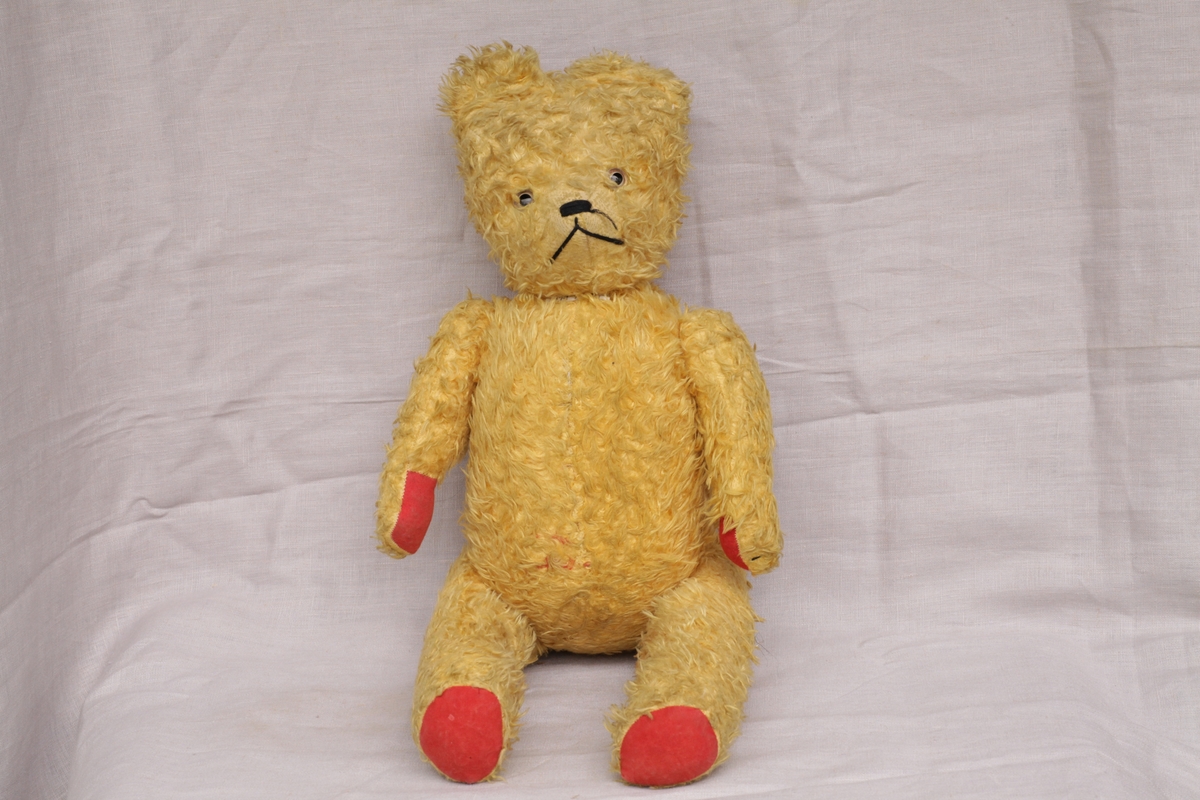 Gul teddybjørn med rød filt på hender og føtter. Øyne av plast og brodert snute og munn. Velbrukt men god tilstand.