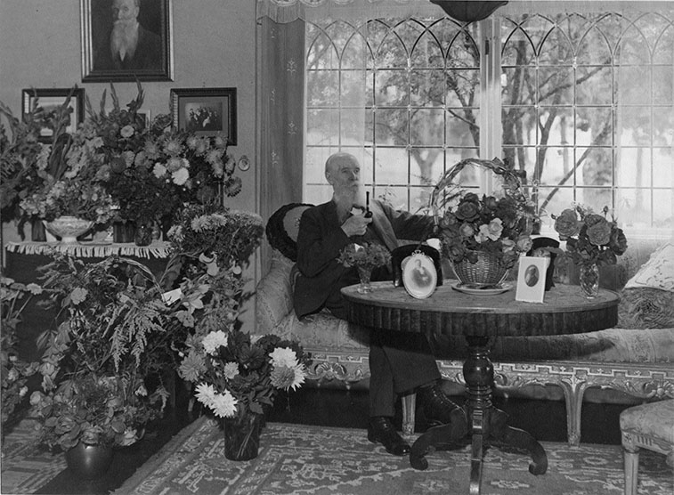 Interiör hos målarmästare G B Berggren på 85-årsdagen på Brahegatan 2-4. Han sitter med en pipa i handen i soffa av äldre snitt med brokadmönstrat tyg. På det runda bordet framför och framförallt på bordet till vänster står det mycket blommor.