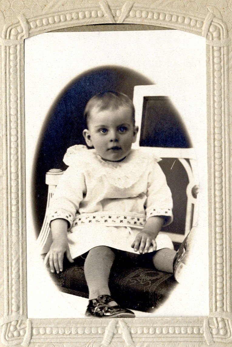 Foto av en liten pojke (?), klädd i ljus kolt med brett skärp. 
Helfigur, en face. Ateljéfoto.