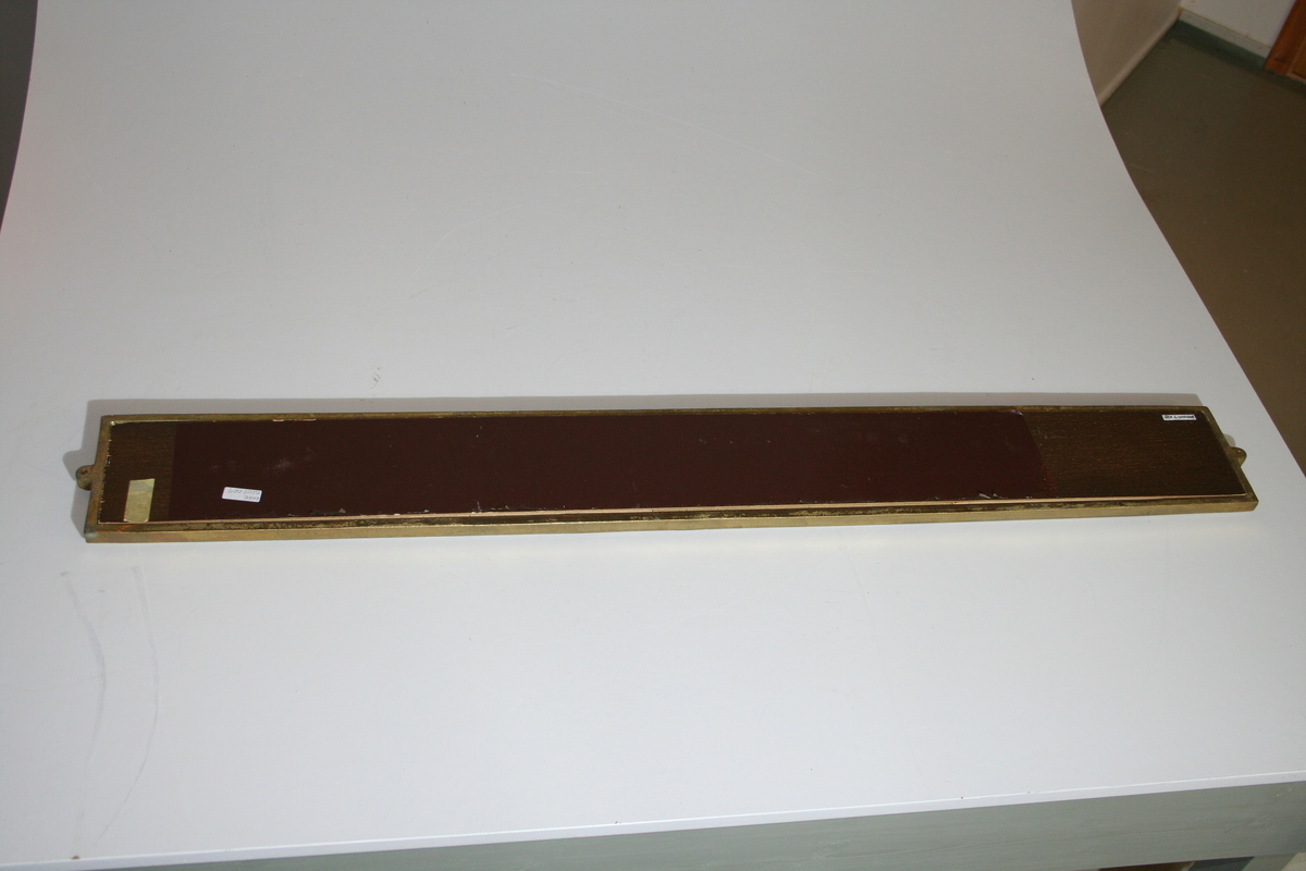 Retangulært, brunt laminatskilt med hvite bokstaver frest ned i platen. Gullmalt metallramme med metalløyer i hver kortside for montering på vegg. På baksiden er det et 75,7 x 10,5 cm bredt felt med folie og en tapebit sitter fast nære den ene kanten.