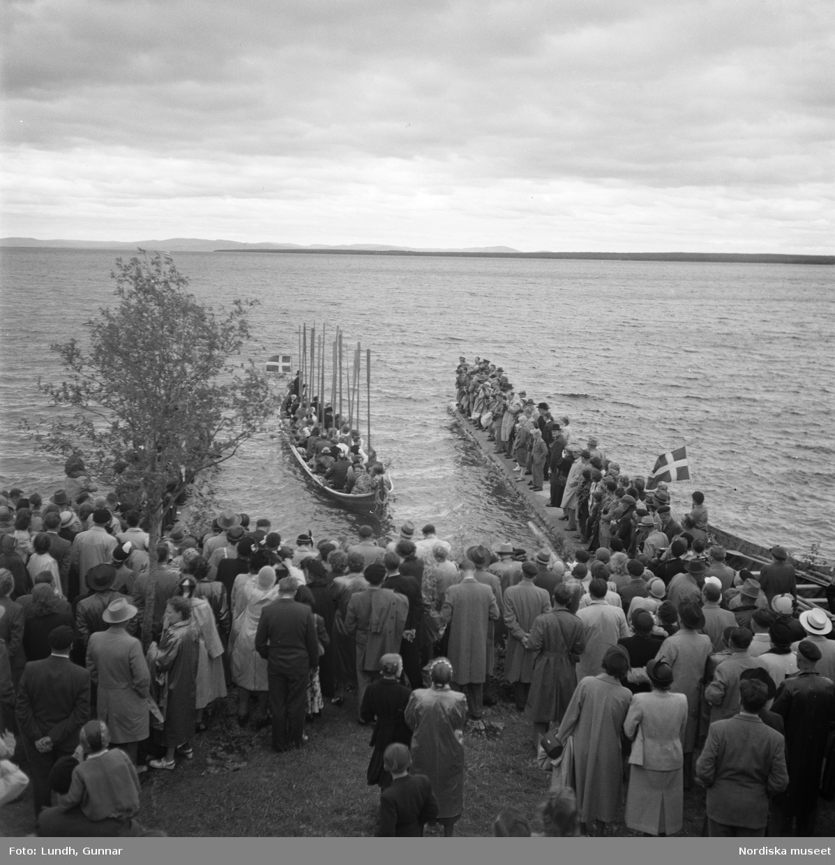 Motiv: (ingen anteckning) ;
En folksamling vid midsommarfirande, en folksamling står på stranden vid en sjö, kvinnor och män i folkdräkt ror en kyrkbåt.