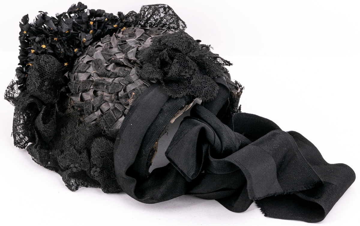 Kapotthatt, svart flätad med tagel och spånband, prydd med spetsar och blombukett, svart med förgyllda ståndare. Svarta knytband av rips.
