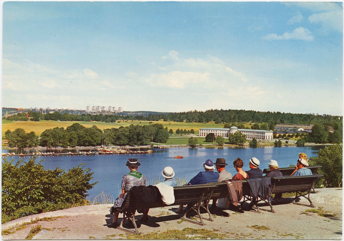 Vykort med motiv från Skansen. "Utsikt över Djurgårdsbrunnsviken."  Skansenbesökare sitter på bänk vid utsiktsplats. Sjöhistoriska museet syns på andra sidan vattnet.