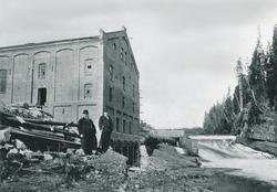 To menn står foran en industribygning i teglstein. I bakgrun