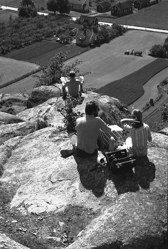 En man och en kvinna har picknick på Grännaberget, på en avsats nedanför Brahehus. En nivå längre ner sitter en man och läser en tidning. Det är varmt och det är sommaren. Längre ner syns Toragården i Uppgränna.