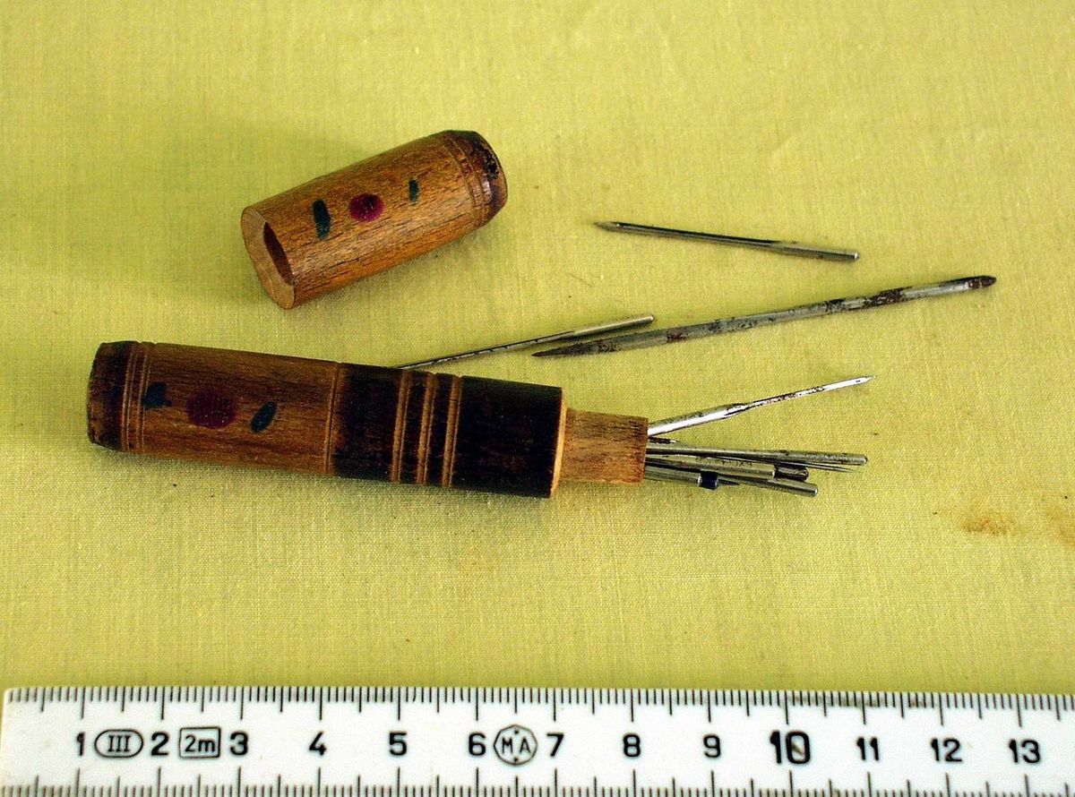 Trehylster for oppbevaring av synåler. Etuiet innheholder flere nåler.
Dekorert med dreide striper, røde prikker og korte grønne streker.