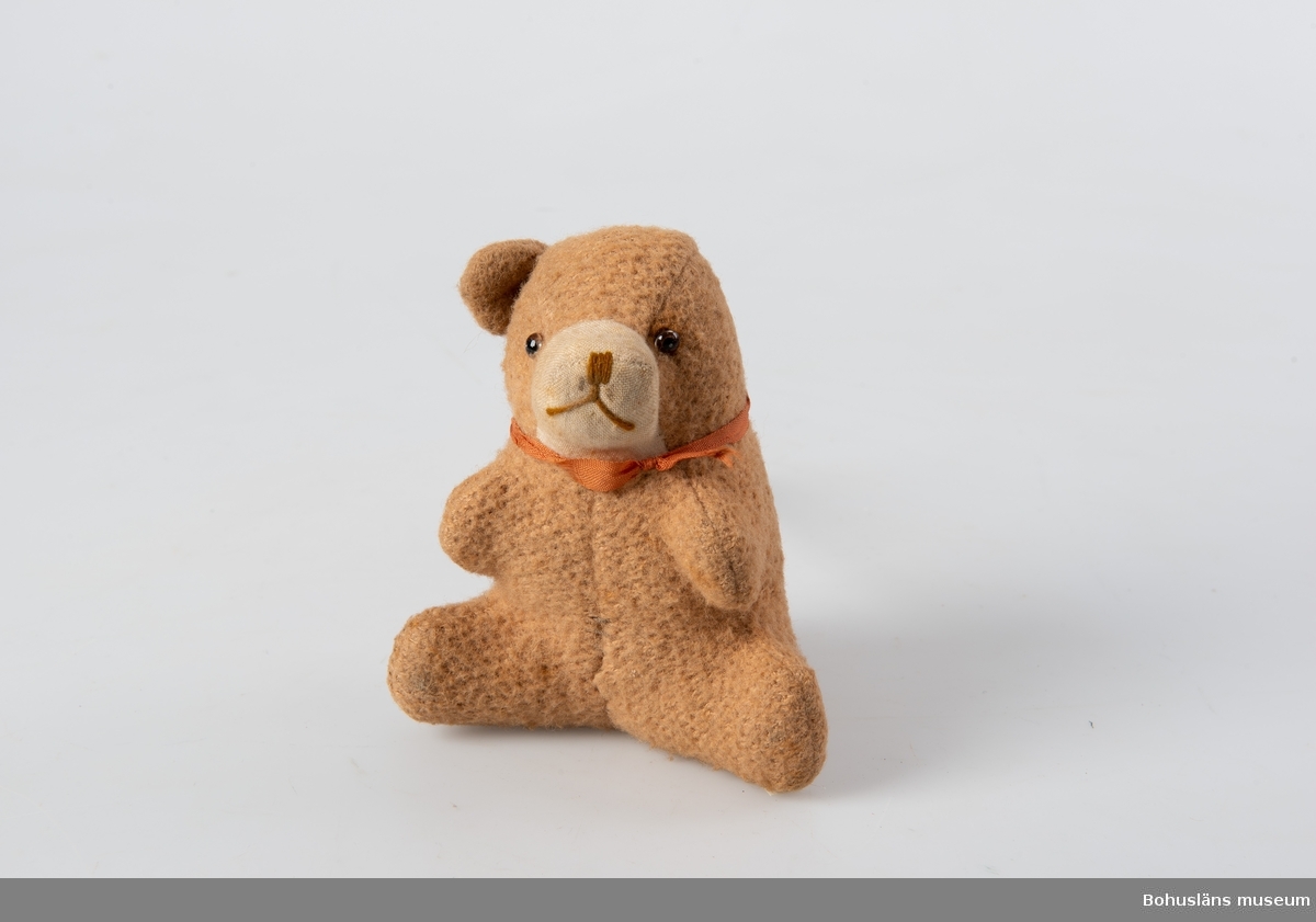 Sittande björn i ljusbrunt bomullstyg, nosparti av ljusgrått linne och nos markerad med brun tråd. Rött band kring hals, ögon av glas, stoppning av krollsplint? Vänster öra saknas.
Personuppgifter se UM026024