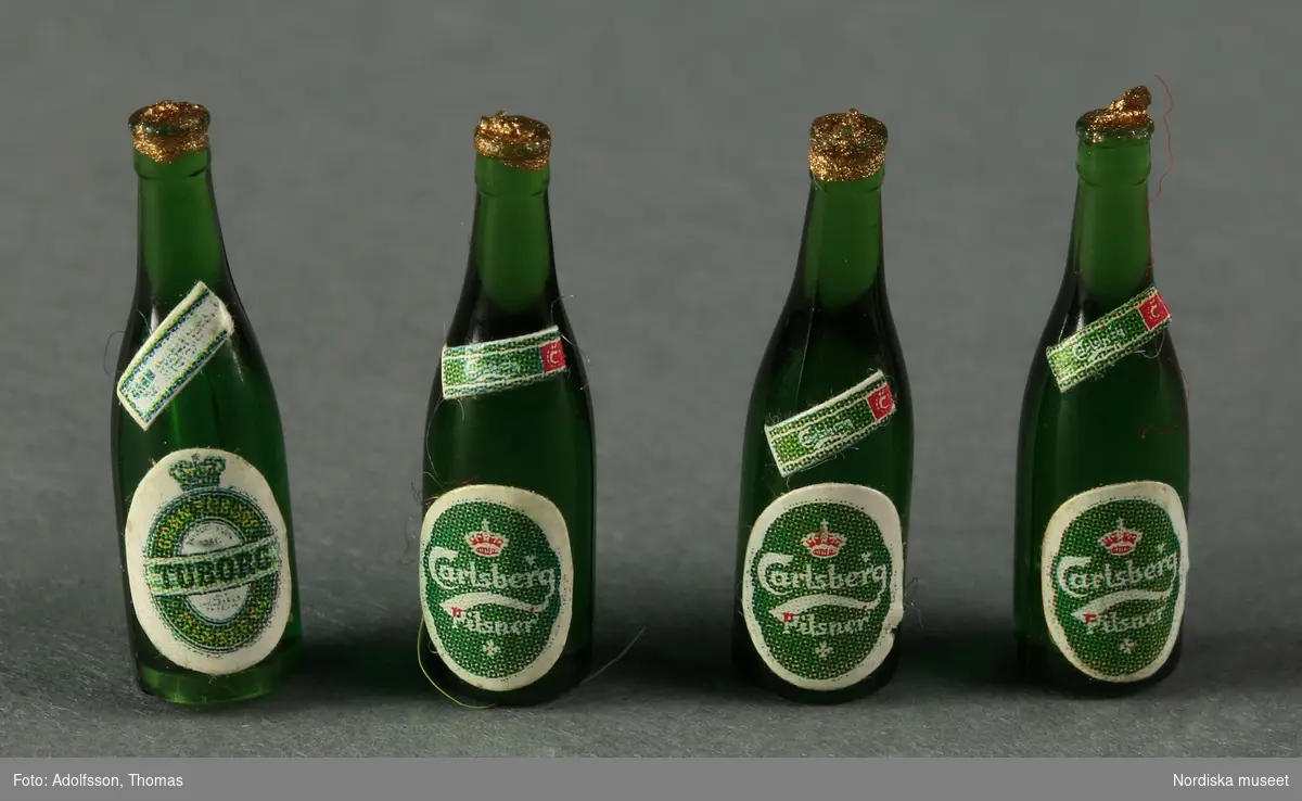 Fyra gröna ölflaskor av plast med etikett av papper. Avbildar dansk pilsner från öltillverkarna a) Tuborg (1 st.) och b-d) Carlsberg (3 st.). Hör till dockskåpsinredningen i matkällaren till dockskåp NM..