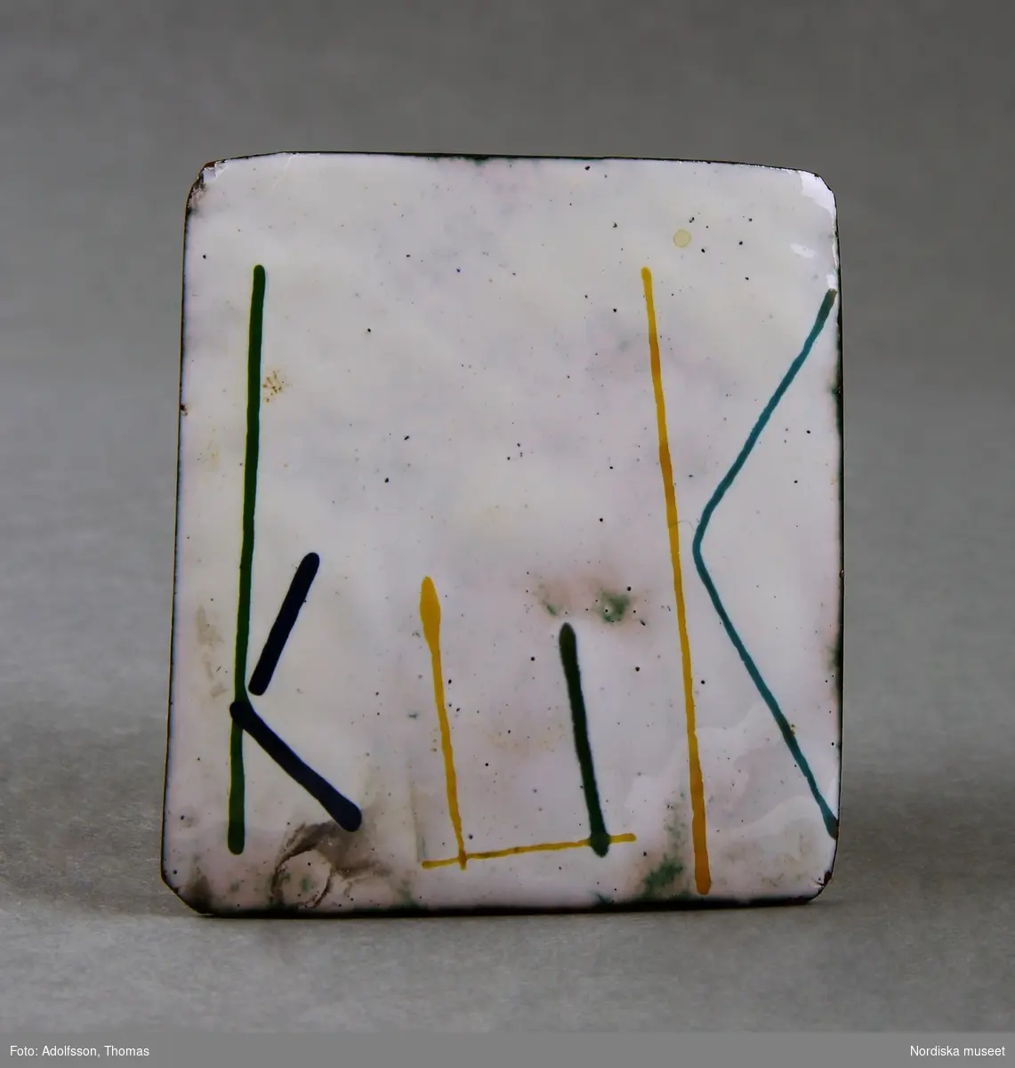Kvadratisk brosch i metall, på baksidan fastlödd nål med lås. Framsidan emaljerad, vit bottenfärg, bokstäver i gult, blått och grönt bildande ordet "KUK".
/Fiffi Myrström, 2014-01-17.