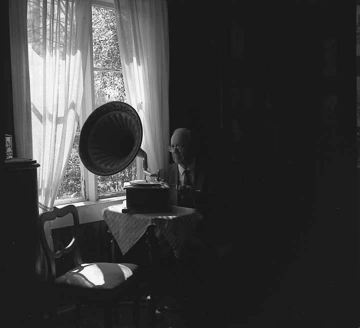 Harry Linder sitter vid ett bord. Han vevar upp en trattgrammofon som står på bordet. Det är mörkt i rummet och genom fönstrets tunna gardiner anas ett starkt solsken.