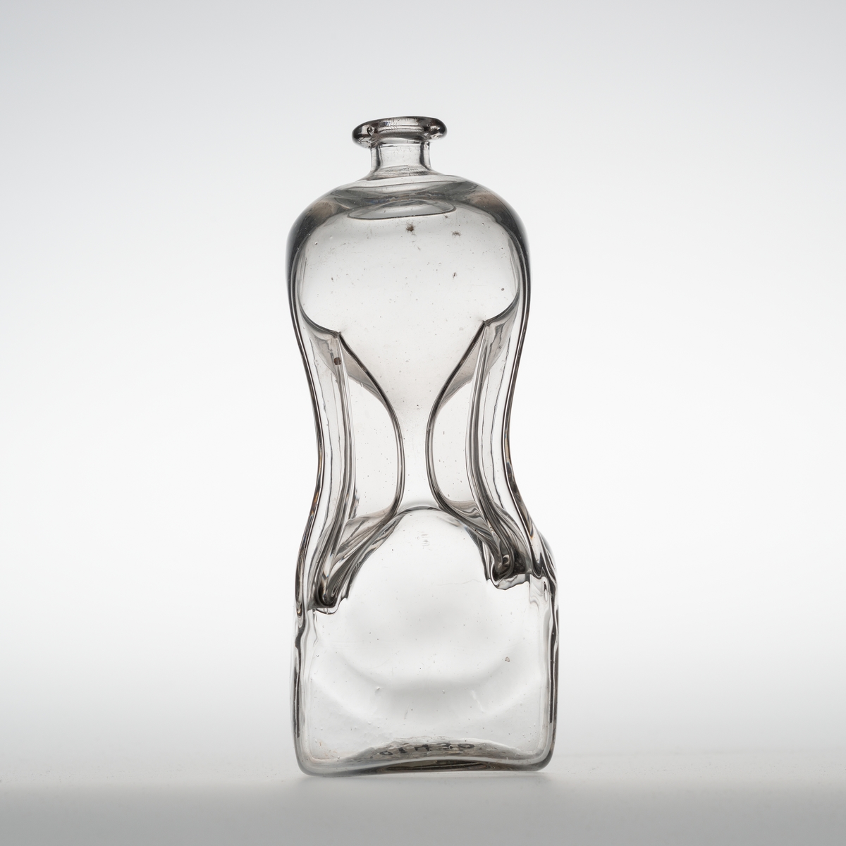 Klunkeflaske i glass, firkantet med sterkt innsvunget, timeglassformet, midtparti og avrundt topp. "Klump" i bunnen etter produksjonen gjør at flasken står ustødig.