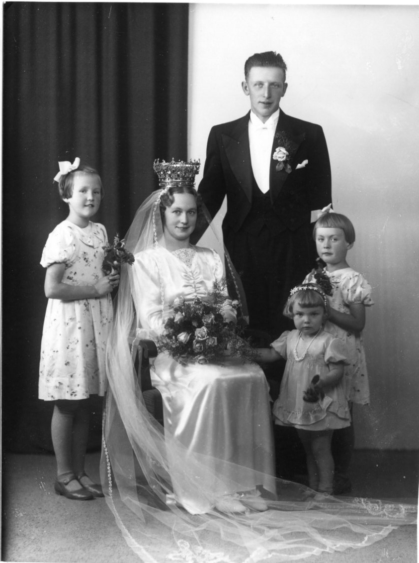 Gruppbild med ett brudpar och tre små flickor - tärnor - som är brudens systerdöttrar. Bruden sitter på en stol med en brudkrona på huvudet och en stor blomsterbukett i händerna. Brudgummen i frack står vid hennes sida.
