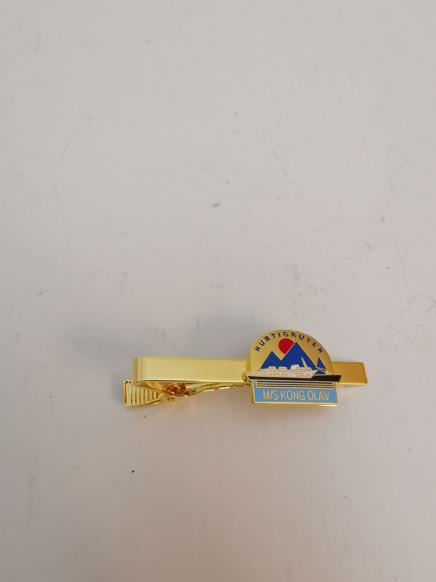 Gullfarget slipsnål med bilde av M/S "Kong Olav" foran på nålen. Nålen har en klype på baksiden for å feste til slips.
