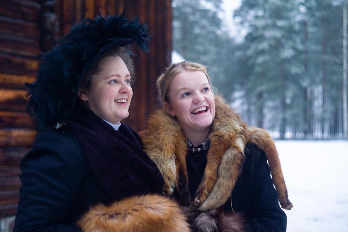 Fotografi av to unge kvinner under Jul i stuene. Den ene kvinnen er dagens husfrue. Hun er kledd i svart, med en fantastisk hatt full av svarte fjær. Hatten sitter litt på skakk. Den andre kvinnen skal spille husfruens søster. Rundt halsen har hun en revepels. De smiler ut mot tunet. Det er snø på bakken.
