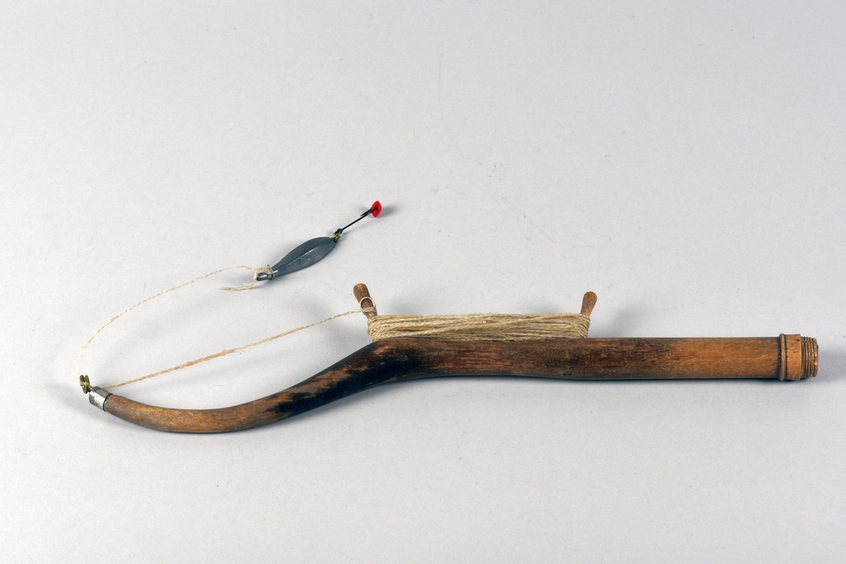 Pimpelspö av trä med lina, drag och krok. Draget är gjort i bly och på kroken sitter en liten röd tygbit. Spöet avslutas med en gängning.