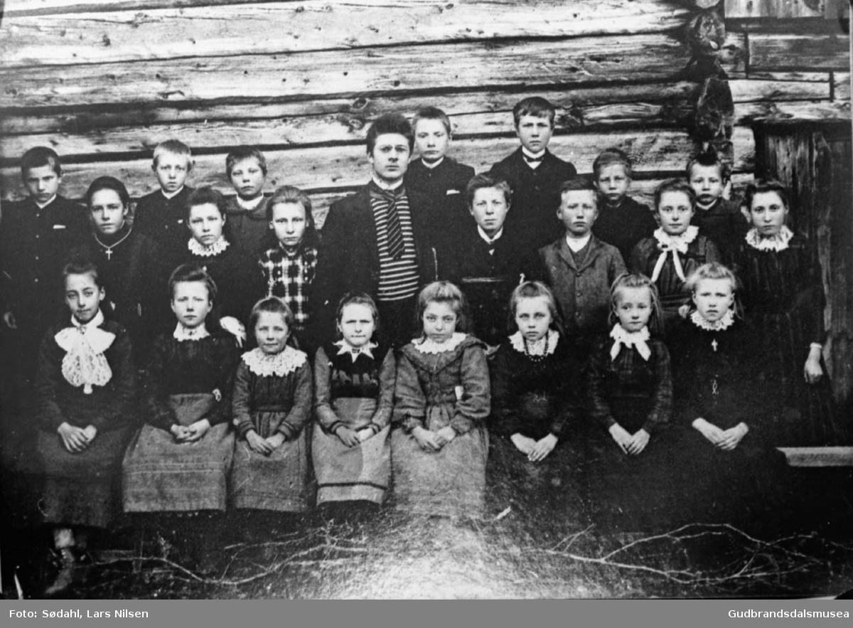 Gruppebilde, stor gruppe av skolebarn og en voksen samlet i husveggen i Uppistugu Bjølverud.

