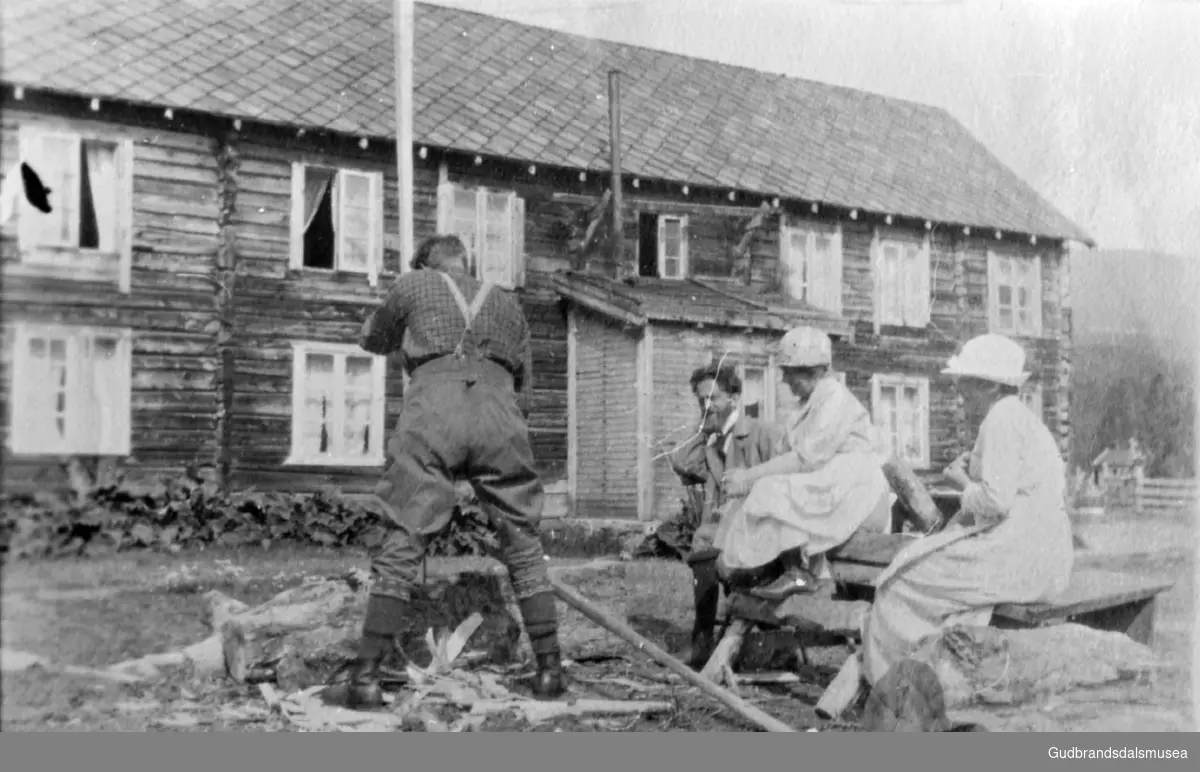 Turister på gårdsplassen i Brygga. En mann, Oddmund?, kvesser stang med øks, to kvinner og en mann sitter og ser på, bygning i bakgrunnen. 