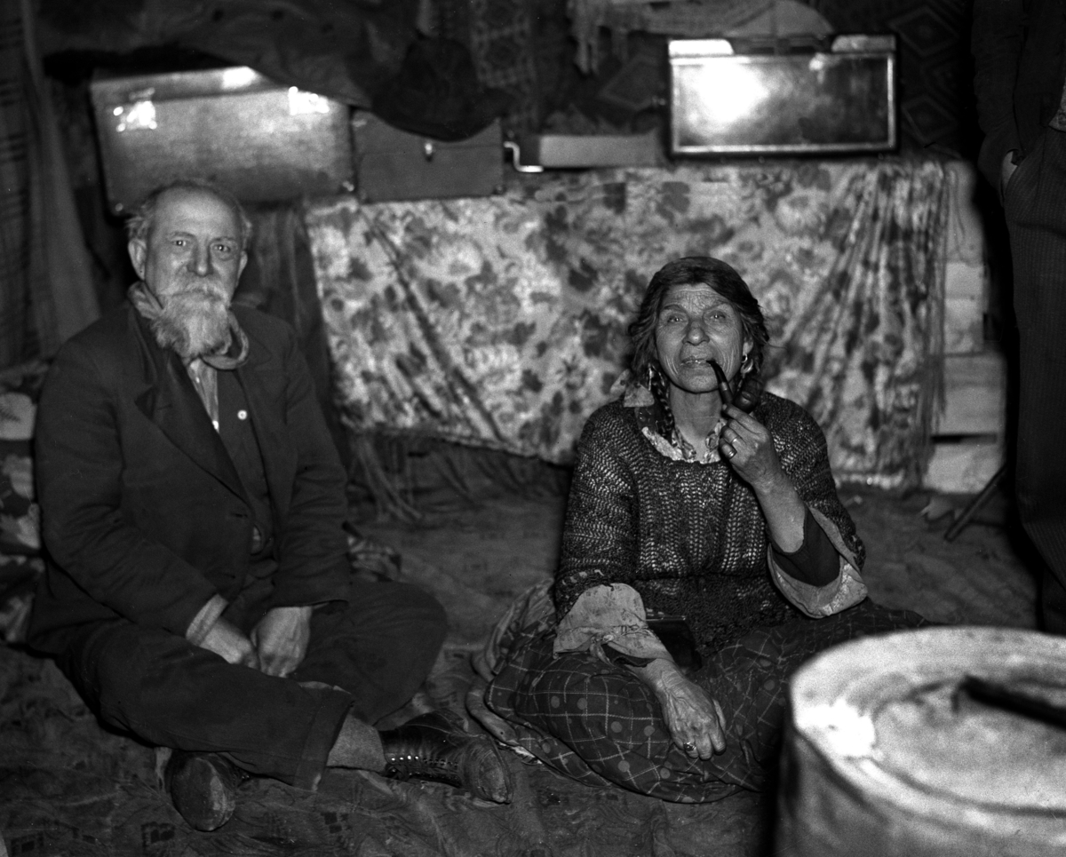 En romsk man och en kvinna sitter på golvet i sin bostad. Kvinnan röker tobak från en pipa.