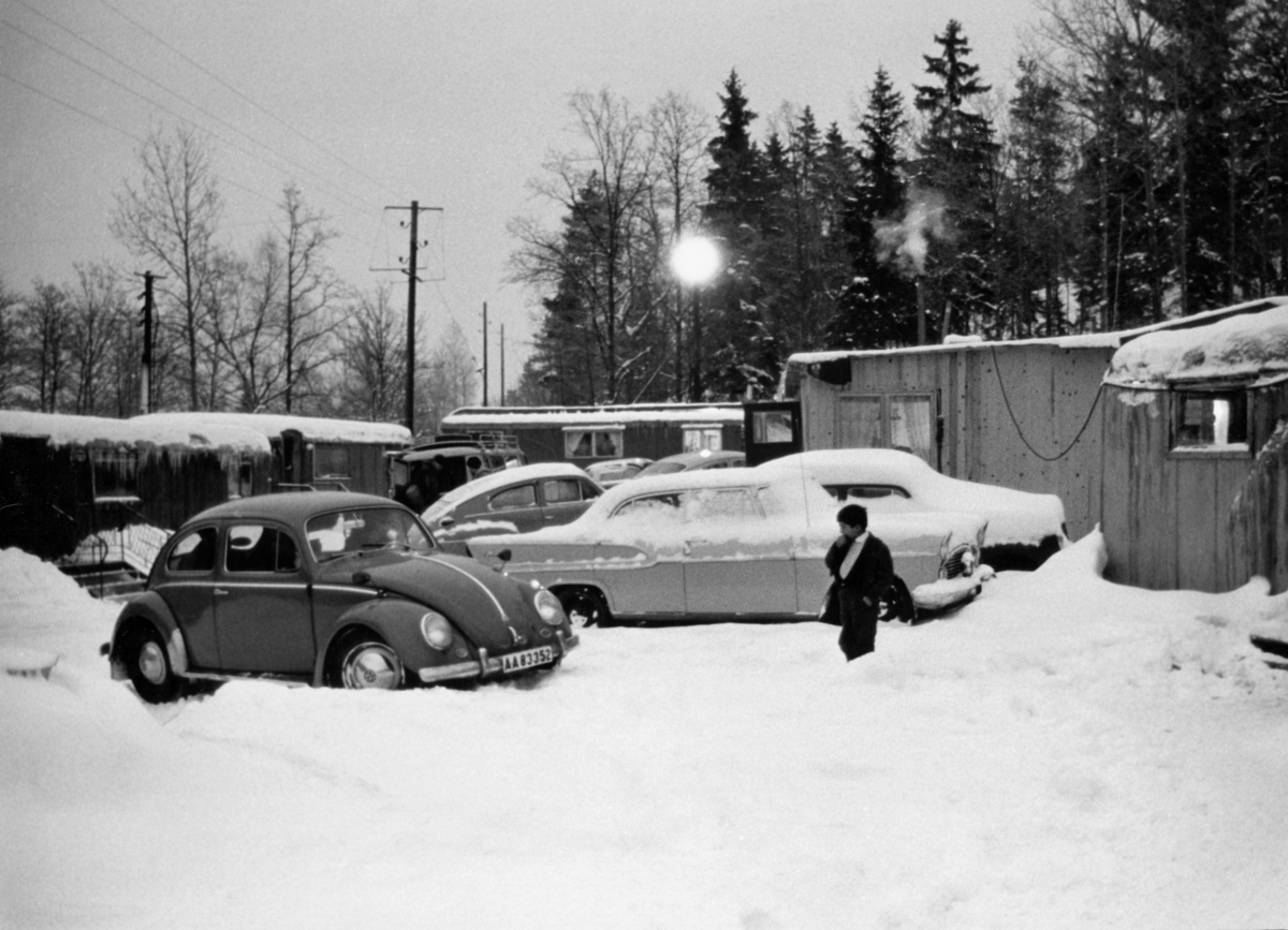 På bilden syns bilar parkerade framför en samling bostadsvagnar. Bilden förmodligen tagen utanför ett romskt läger i Stockholm 1963.