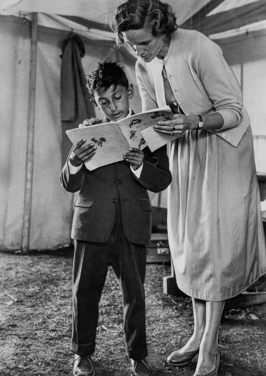 En romsk pojke läser högt ur en bok på uppmaning av sin lärarinna som står intill honom. Händelsen tar plats under en sommarskola i ett romskt läger, 1957 i Älvsjö.