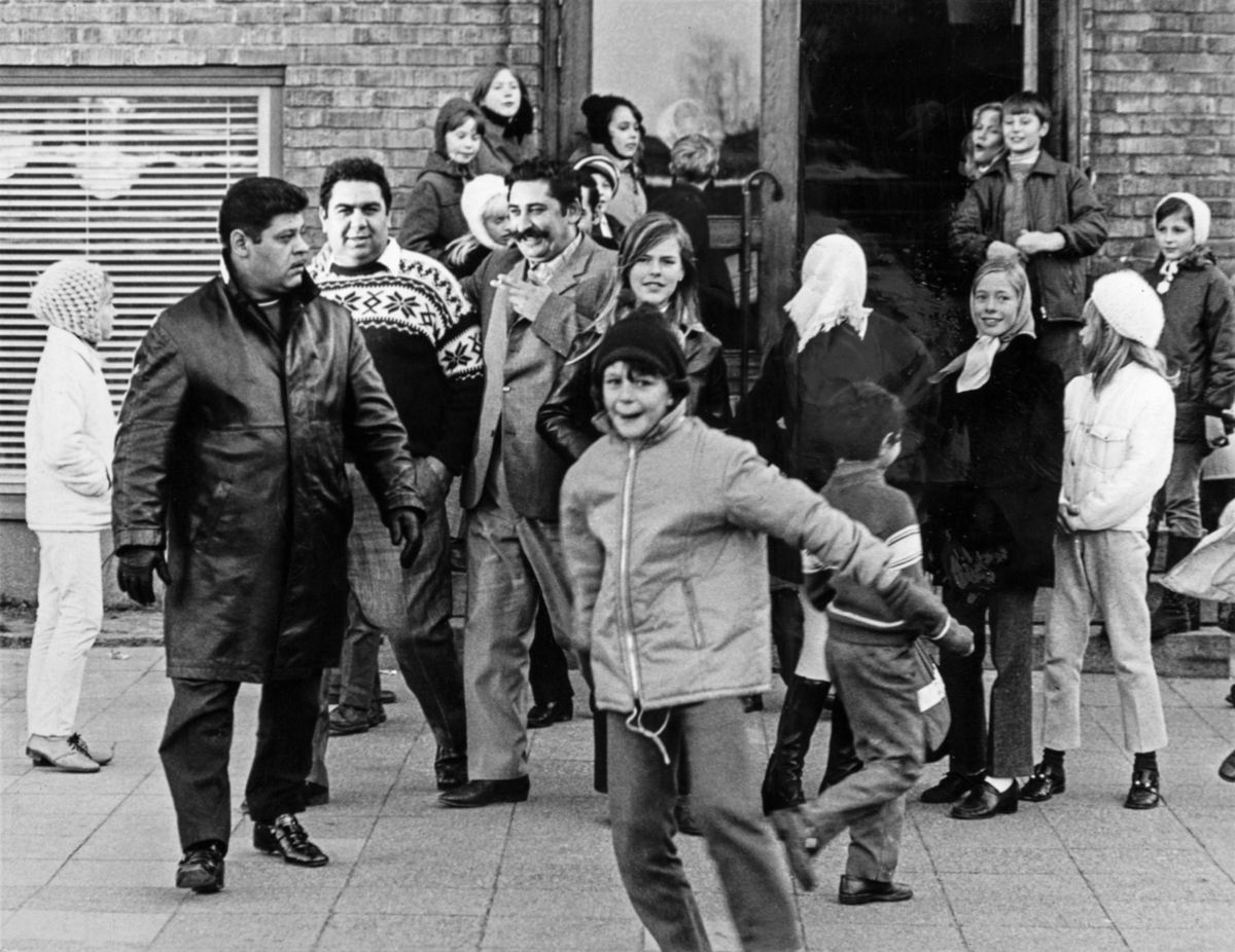 Utanför en skola i Malmö 1969 är en grupp vuxna och barn samlade. Några barn springer och några tittar mot kameran.