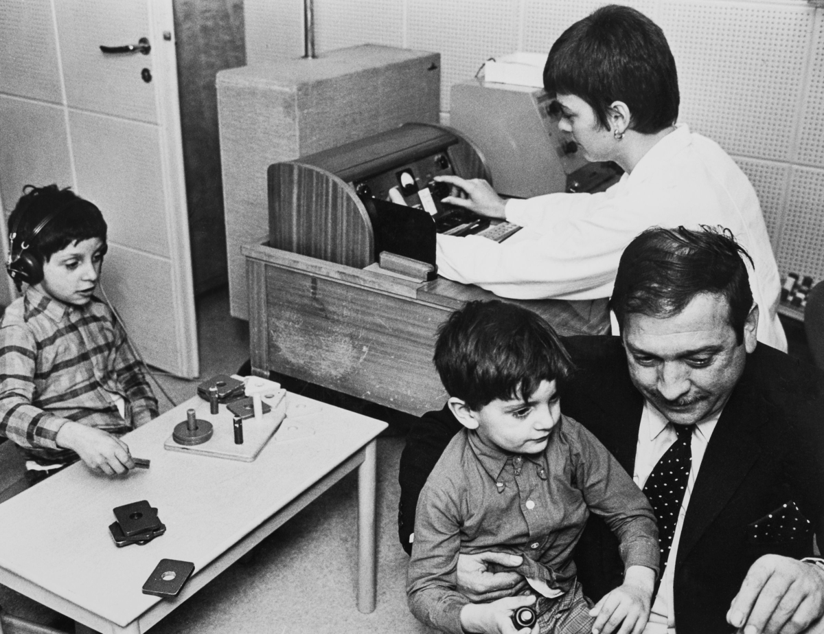 Två romska pojkar undersöks vid Karolinska sjukhusets hörselklinik. En pojke sitter i knät på en man och en sitter med hörlurar på huvudet.