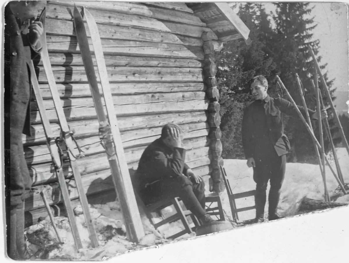En gruppe menn står og sitter utenfor en tømmerbygning. Ski oppetter veggen og staver i snøen.
