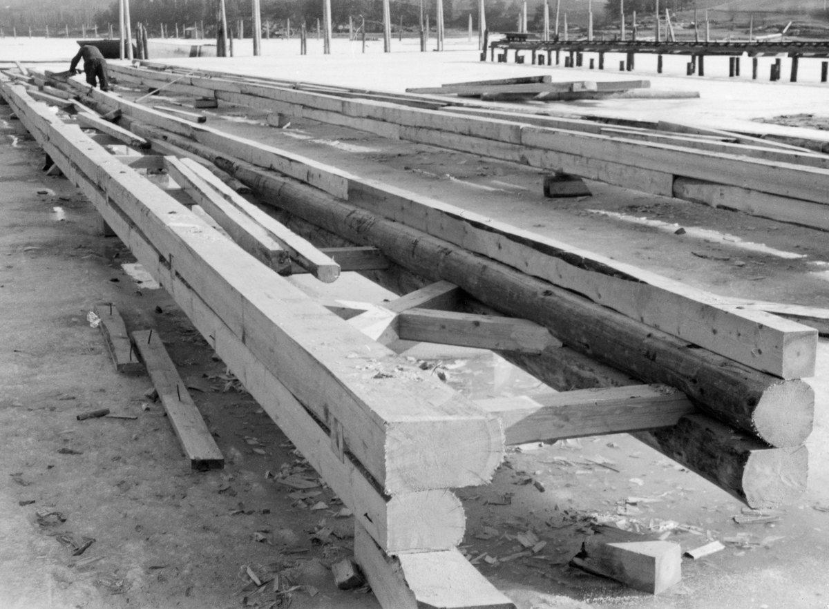 Bygging av nye ganglenser ved lenseanlegget på Eidet i Tune i Østfold vinteren 1953.  Arbeidet ble utført på isen på innsjøen Visterflo, ved sjølve lenseanlegget, der disse konstruksjonene skulle brukes.  Rammekonstruksjonen, som det ble arbeidet med her, besto av tømmer som var rettskåret på tre kanter og boltet sammen i anslagsvis 20-30 meters lengde.  Rammene var cirka en meter brede, med de nevnte sammenboltete tømmerkonstruksjonene på langsidene og med dels rette, dels krysslagte stag av tre mellom.  Konstruksjonen lå på kabber på isen, og ved den fremre enden av den påbegynte ganglensa som ligger sentralt i bildeflata ser vi en lensearbeider som sagde på en bjelke.  Arbeidsstedet er omgitt av pælerekker som stikker opp gjennom isflata.  