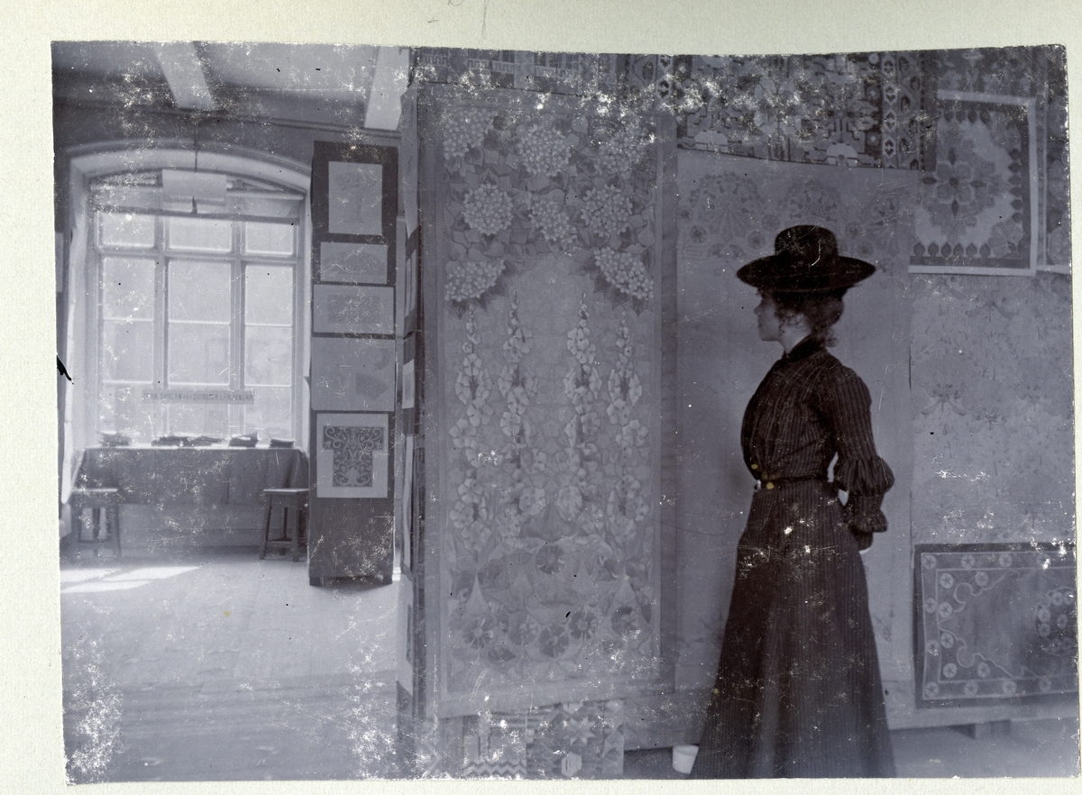 Stockholm. 
Kvinna stående framför vägg med bl.a. blomstermålningar. 1901.