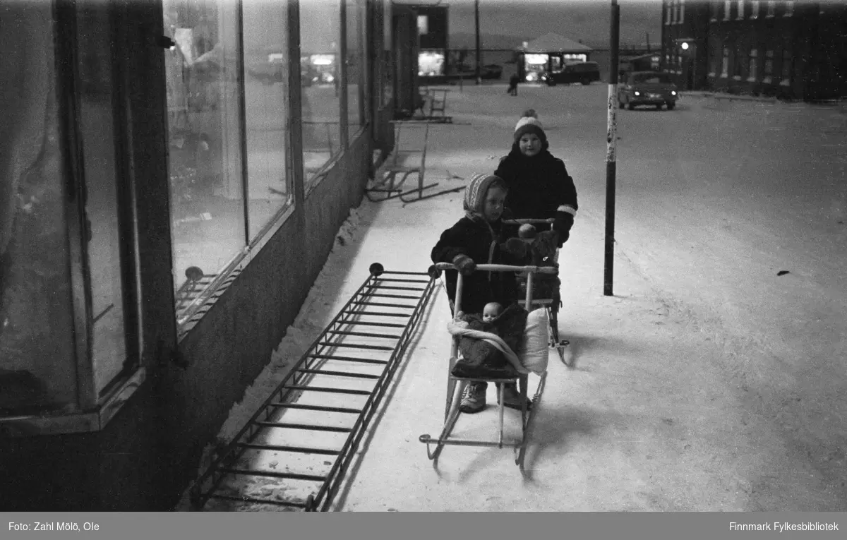 Vadsø, desember 1967. Juletider. To barn med sparkstøtting og dukke på passasjersetet.