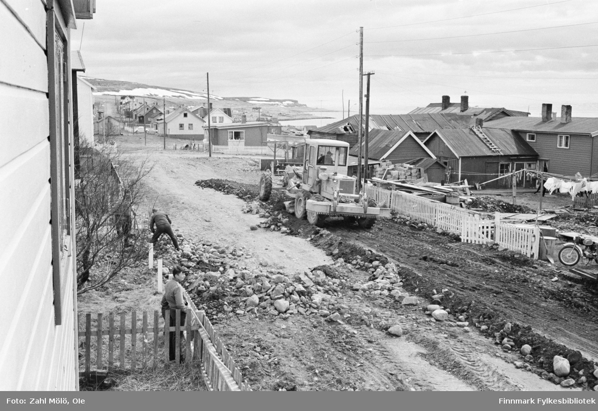 Vadsø 1969. Fotoserie av Ole Zahl Mölö. Veiarbeid og dosering av jord og stein med bulldoser. En mann i arbeid med stein og en gutt som står og bivåner arbeidet.