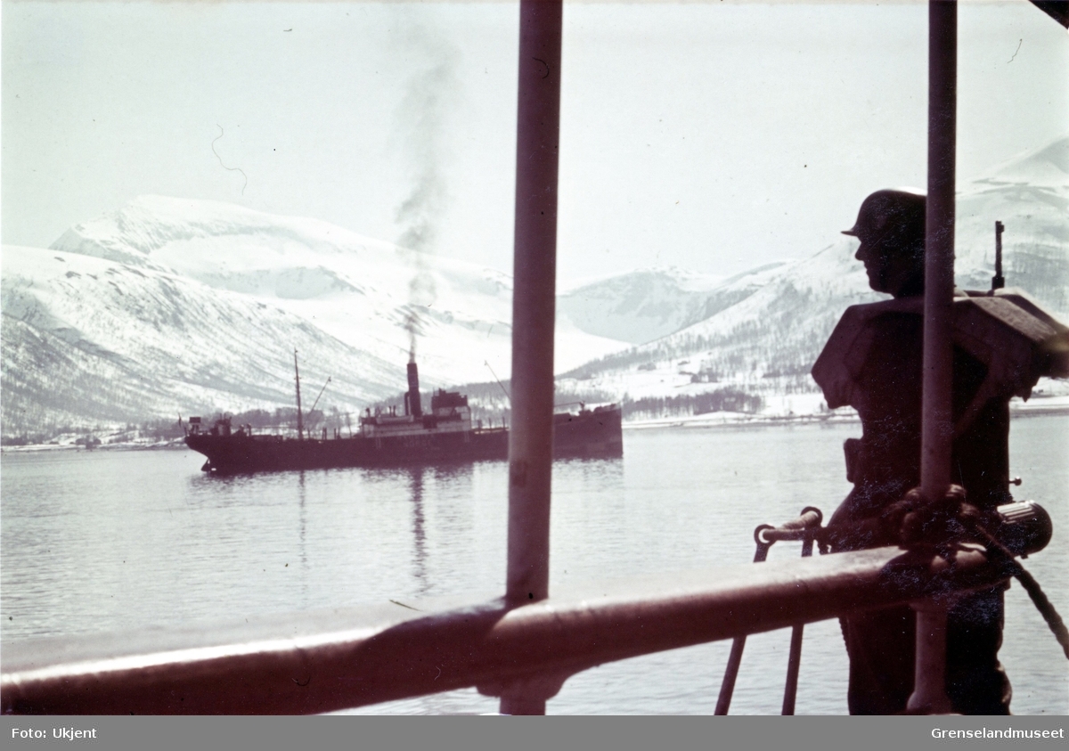 Tromsøysundet med Tromsdalstind i bakgrunnen. Skipet midt i bildet har påskrift NORGE. Navnet er vanskelig å lese. Soldat med våpen i forgrunnen til høyre.