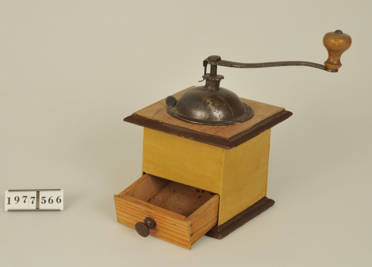 
Kaffekvarn med fyrkantig låda av gulmålad plåt med botten och översida av trä. Skål av plåt samt vev av järn med handtag av trä.