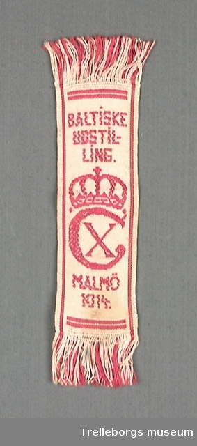 Vitt mönstervävt band med rött mönster och text. Texten lyder: BALTISKE UTSTILLING MALMÖ 1914 Mitt på bandet finns också ett C med ett X innåt och en kungakrona ovanför. 2 cm lång frans i båda ändarna.